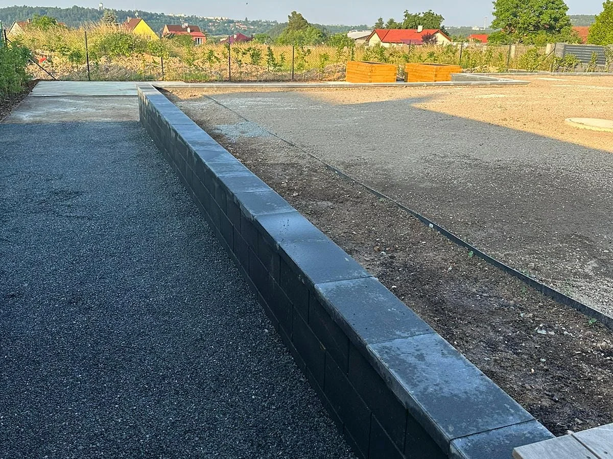 Opěrná zídka z tmavých betonových tvárnic se zákrytovými deskami, vedle plocha z kamenné drtě
