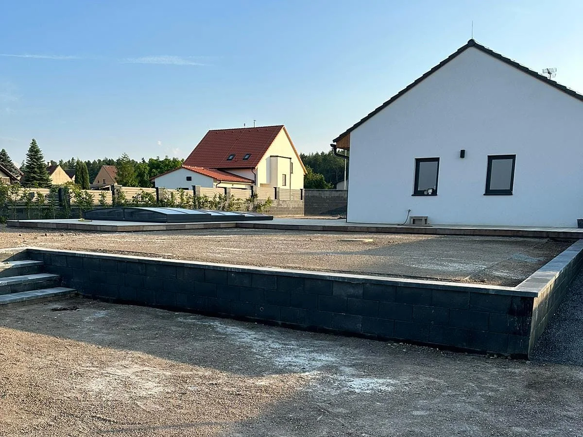 Opěrná zídka se schody, za ní dřevěná terasa se zastřešeným bazénem a nový rodinný dům